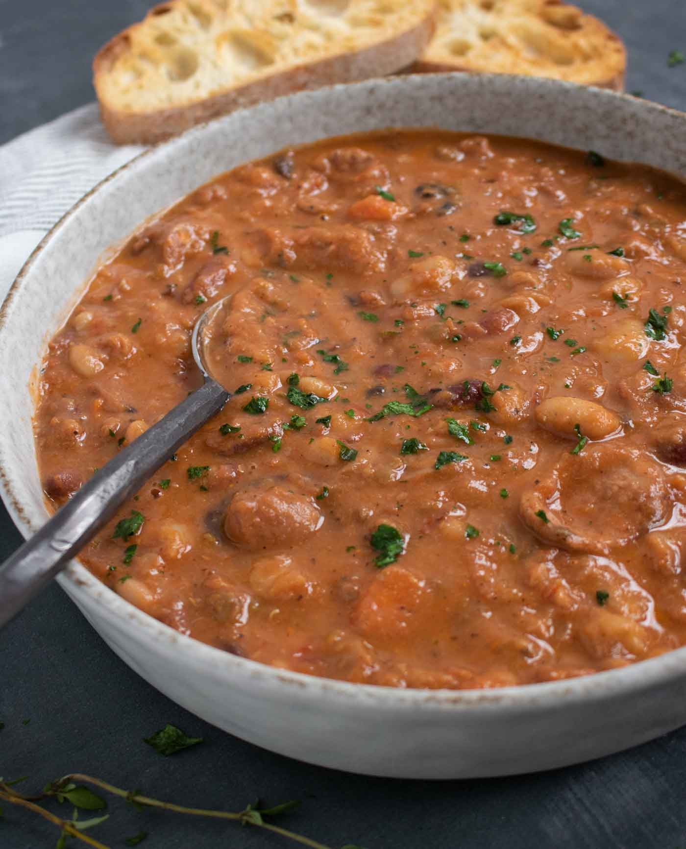 A side-angled view of a bowl of 15 Bean Soup with a spoon, and bread slices in the background.