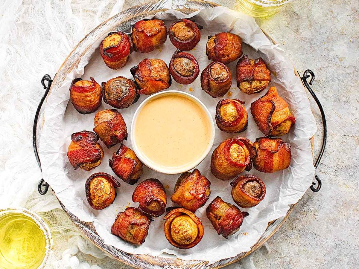 Bacon wrapped potato appetizers on a white serving tray with dip.