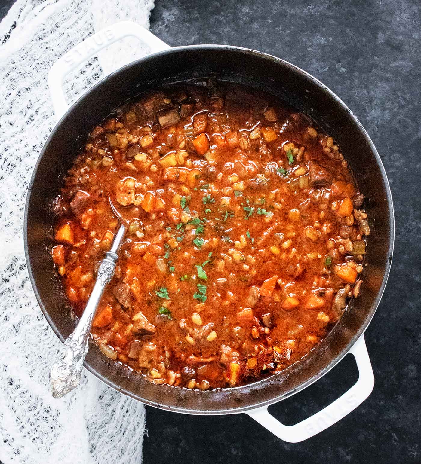 Overhead view of Beef Barley Soup in a large white Dutch oven with a serving spoon.