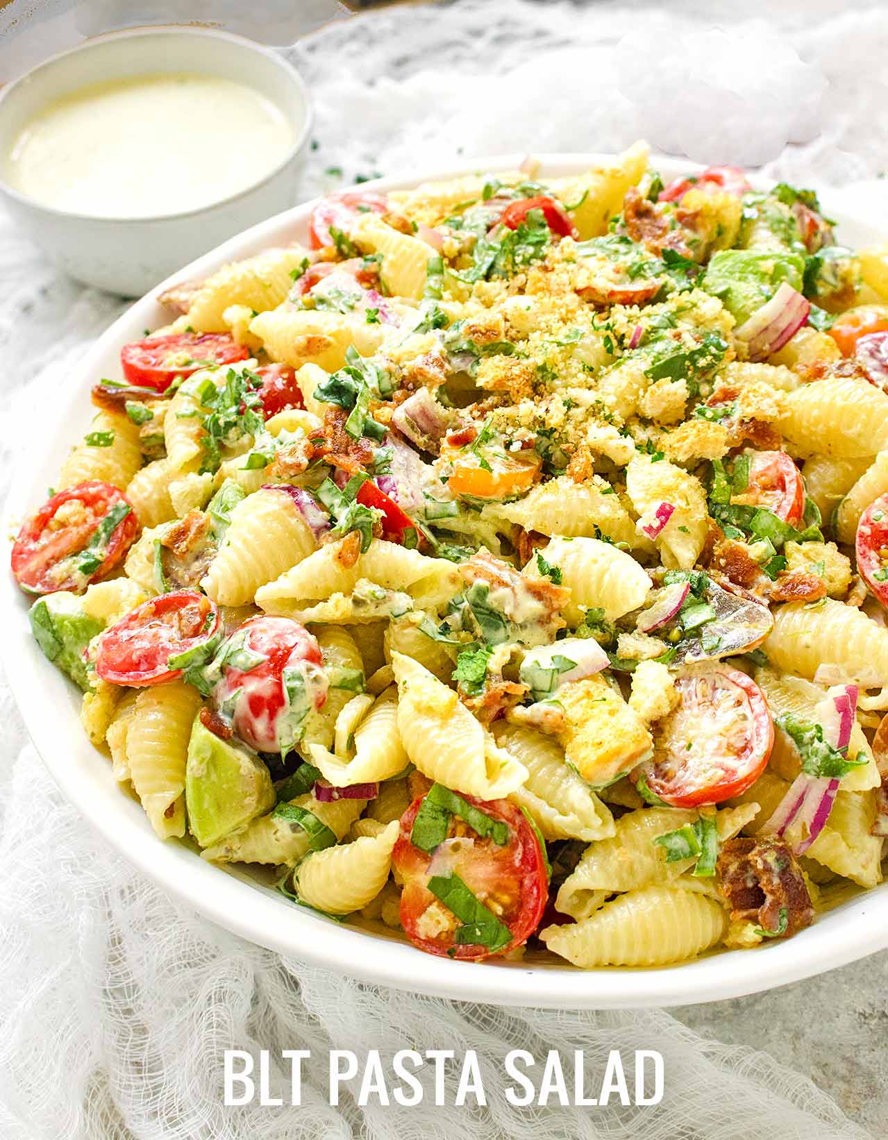 Side view of a big bowl of BLT pasta salad, with a small bowl of extra dressing in the background