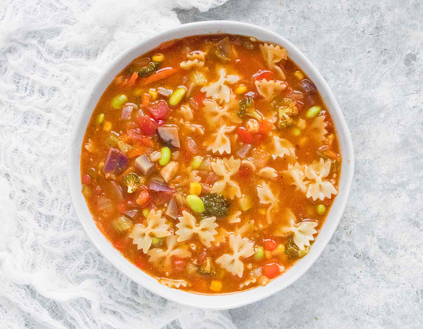 A bowl of vegetable soup with bowtie pasta on a white background
