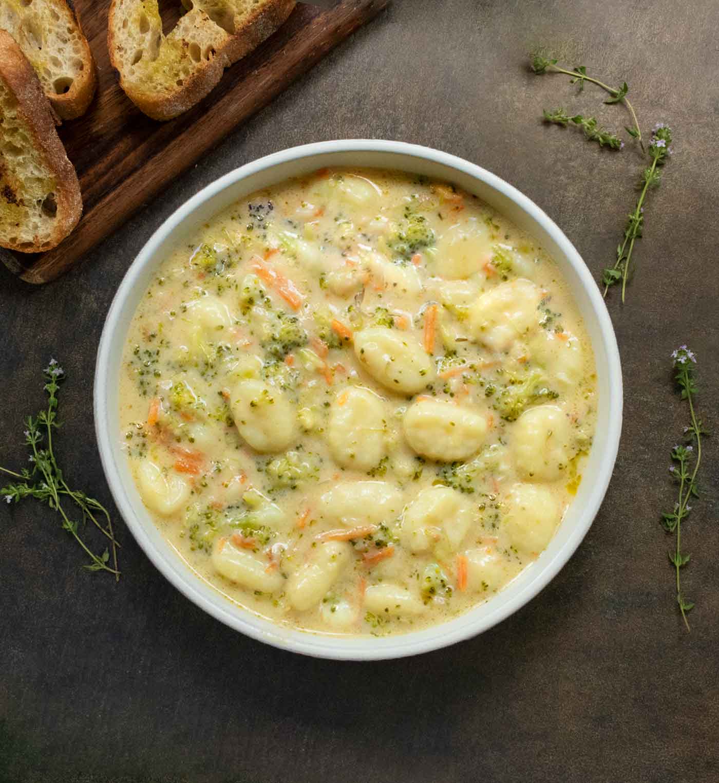 An overhead view of Broccoli Cheddar Gnocchi Soup in a white bowl with a side of baguette slices on a wooden board.