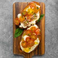 Two slices of Cherry Tomato Bruschetta on a bread board with basil leaves.
