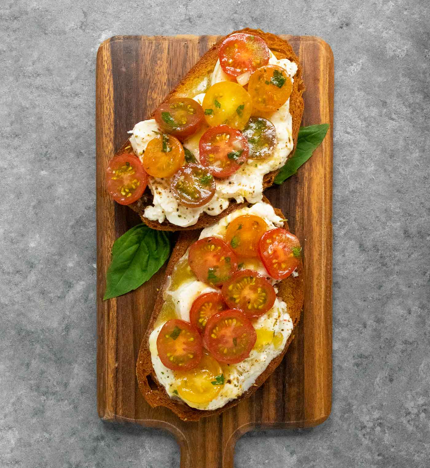 Two slices of Cherry Tomato Bruschetta on a bread board with basil leaves.