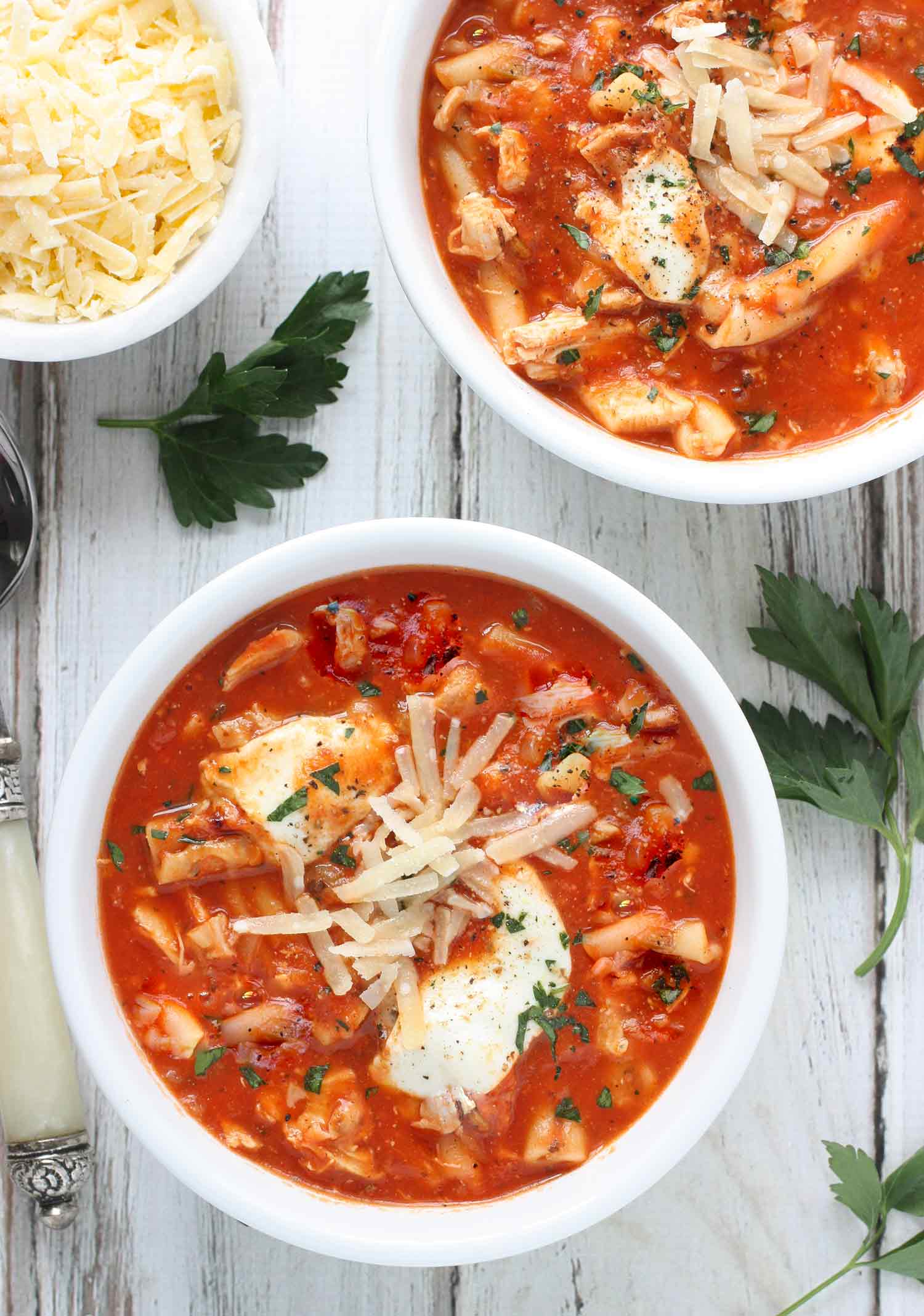 Two bowls of Chicken Parmesan Soup, ready to serve with a side bowl of shredded parmesan cheese.