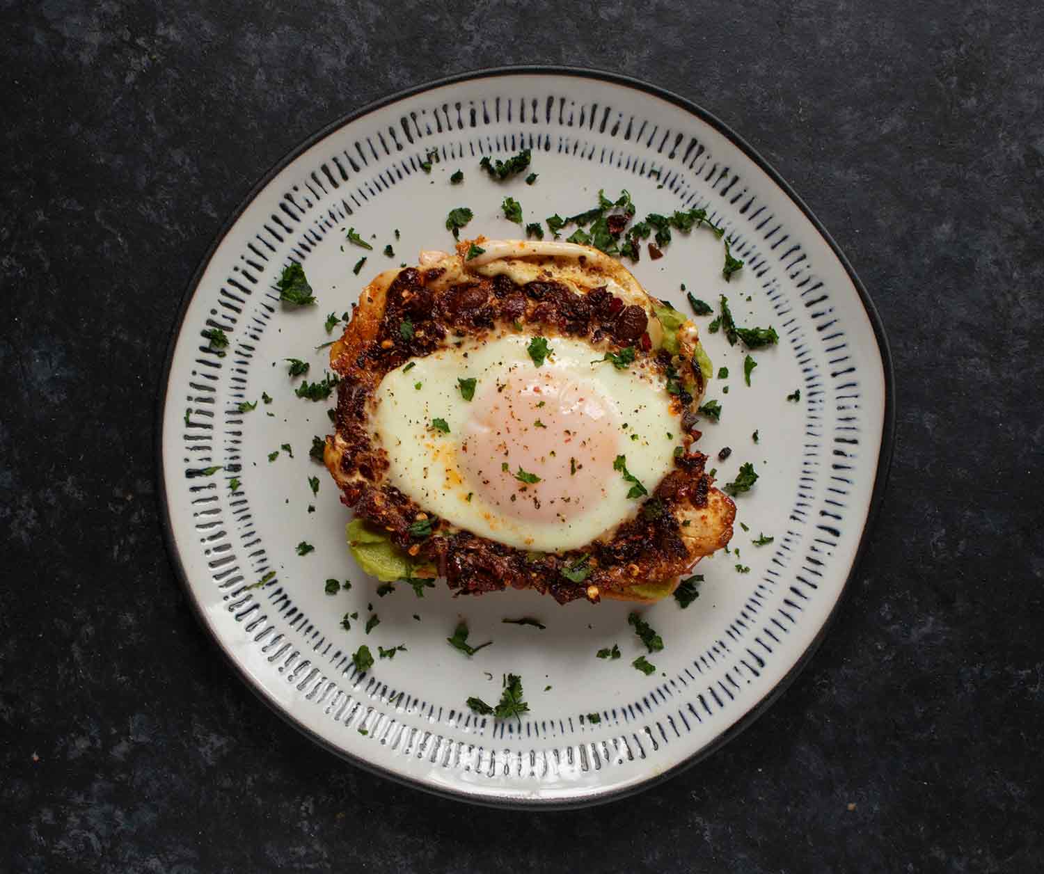A Chili Crisp Fried Egg on avocado toast on a white plate with black rim.