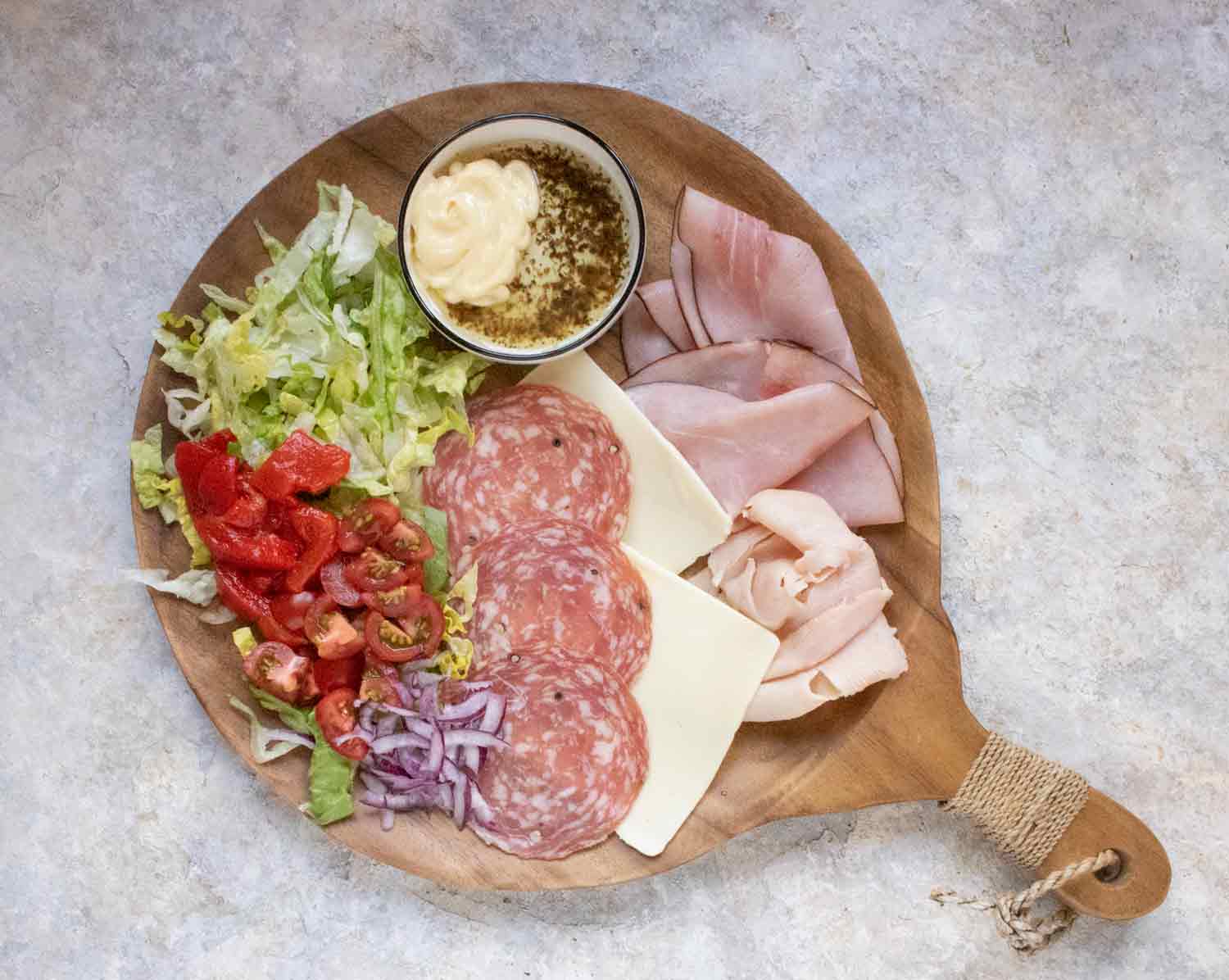 Ingredients for a Chopped Grinder Fried Toast sandwich spread on a wooden cutting board, including ham, turkey, salami, cheese, lettuce, tomatoes, roastsed red peppers, and red onions.