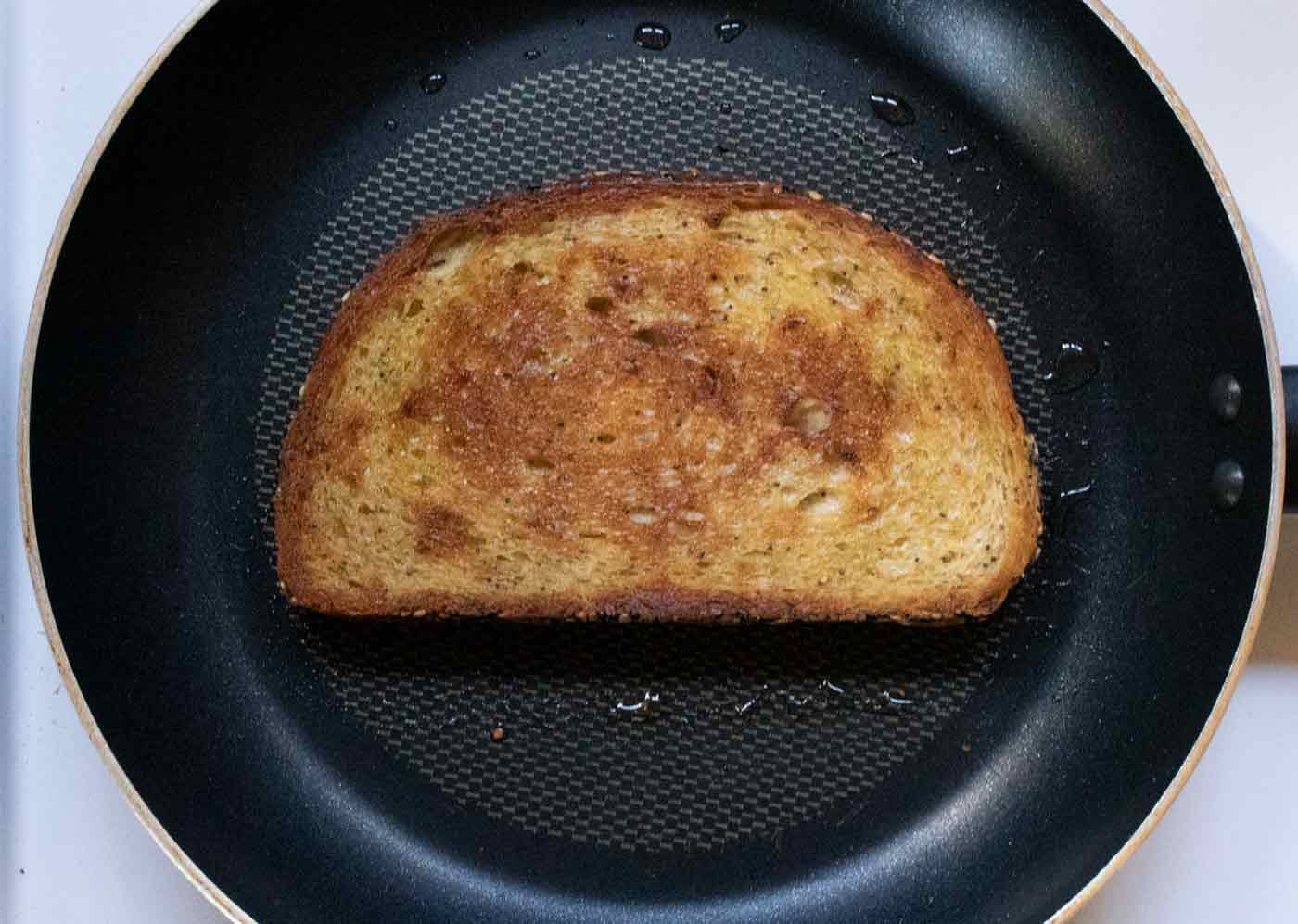 A slice of sourdough bread drizzled with oil toasting in a skillet on the stove.