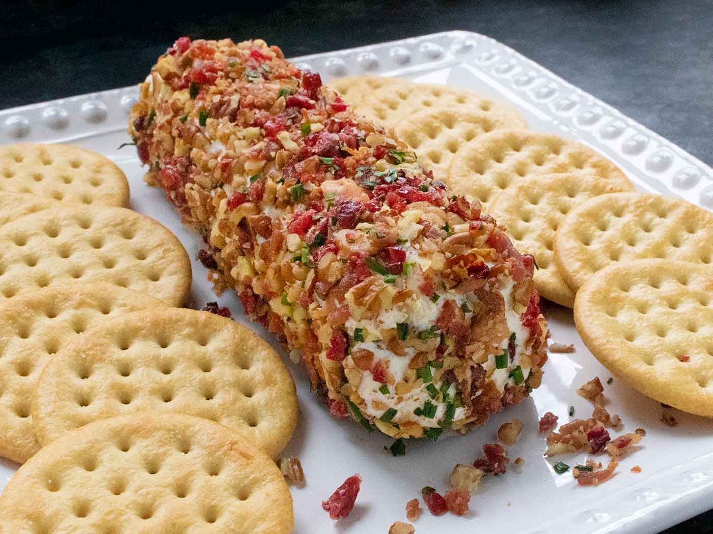 A Cranberry Pecan Bacon Cheese Log on a square white plate with crackers.