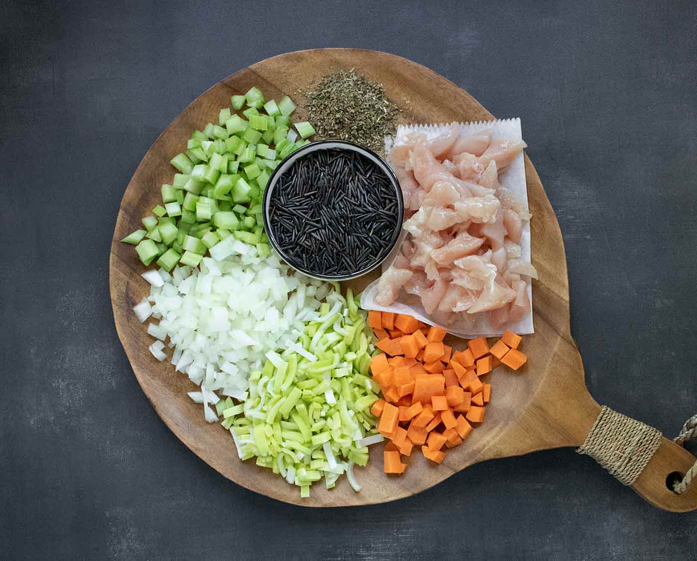 The main ingredients of Creamy Chicken Wild Rice Soup on a wooden serving board. Shown from top, clockwise: poultry seasoning, chicken, carrots, leeks, onions, celery, wild rice.