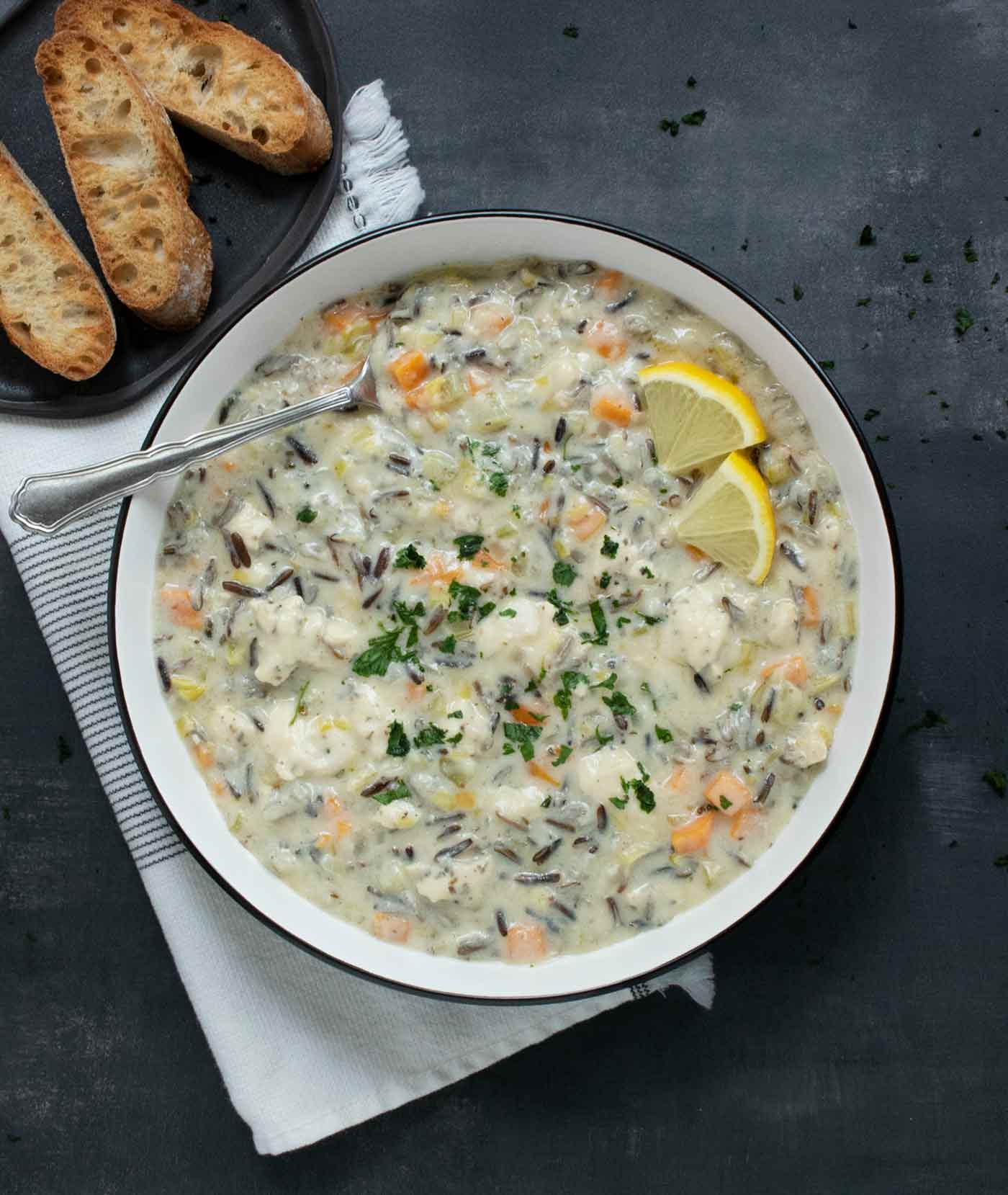 Creamy Chicken Wild Rice soup in a white bowl with a black rim, and a spoon, with a side of sliced bread.