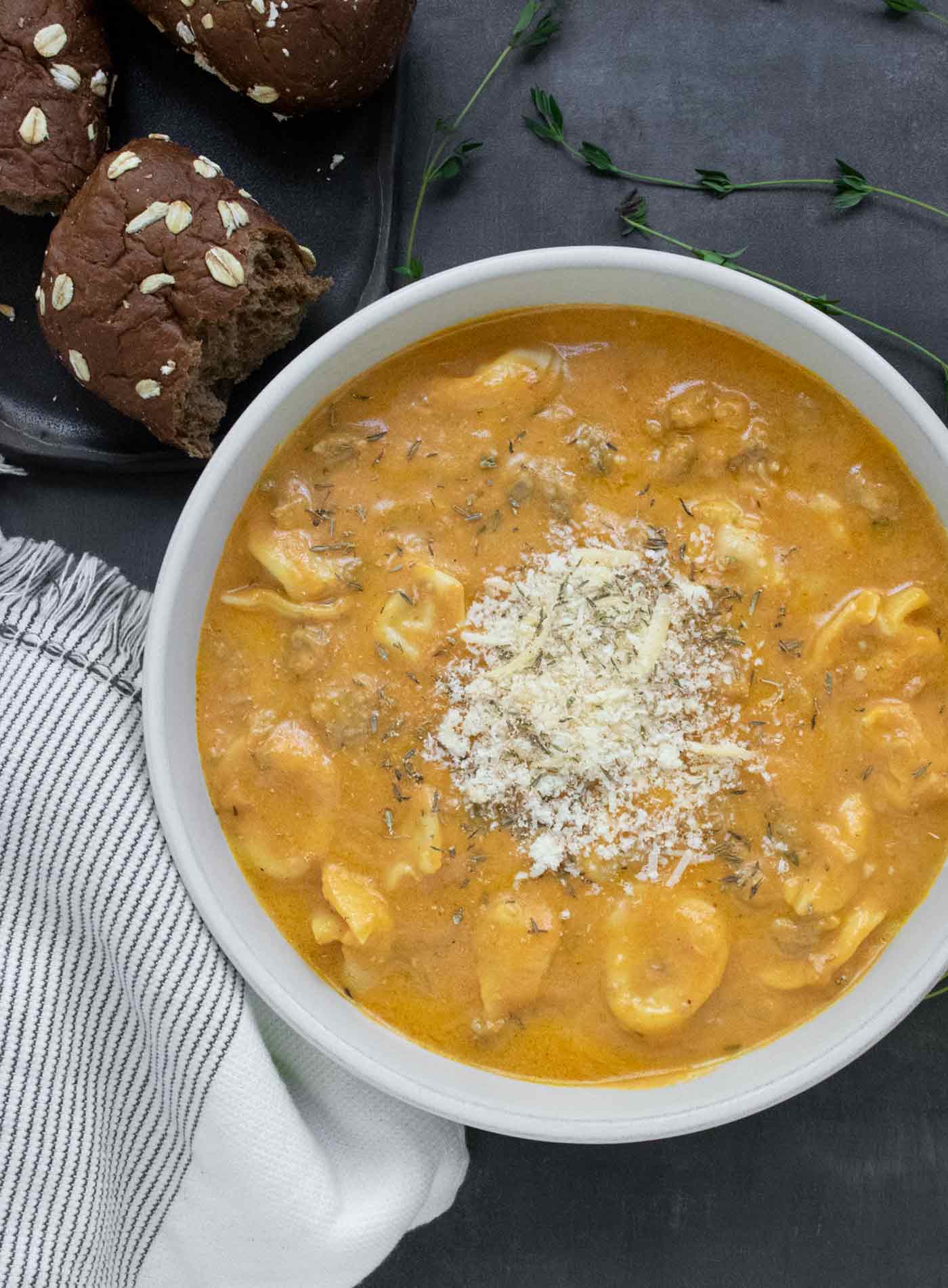 A bowl of Creamy Pumpkin Sausage Tortellini Soup with a side of brown bread dinner rolls.