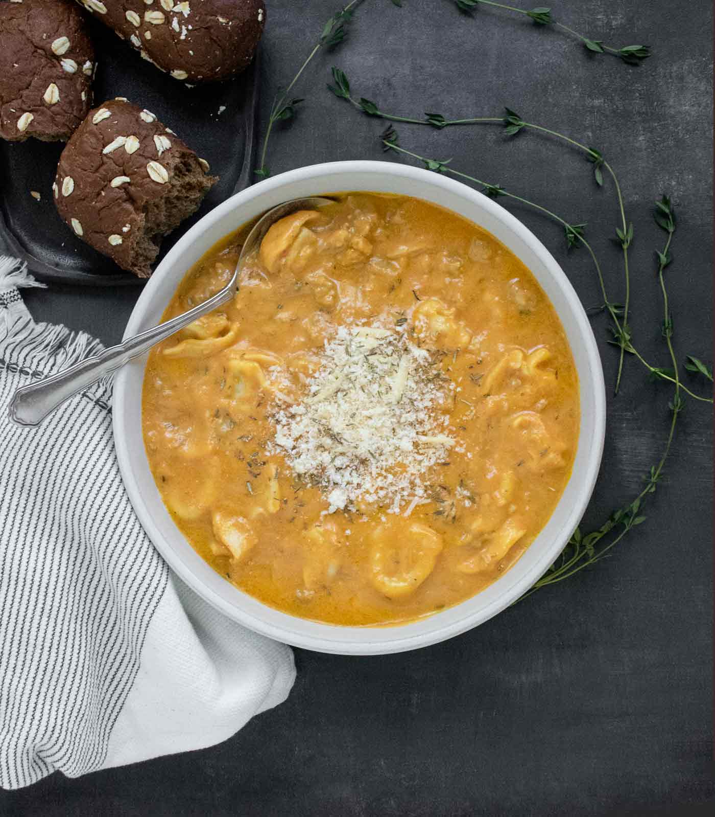 An overhead shot of a bowl of Creamy Pumpkin Sausage Tortellini Soup with a side of dinner rolls.