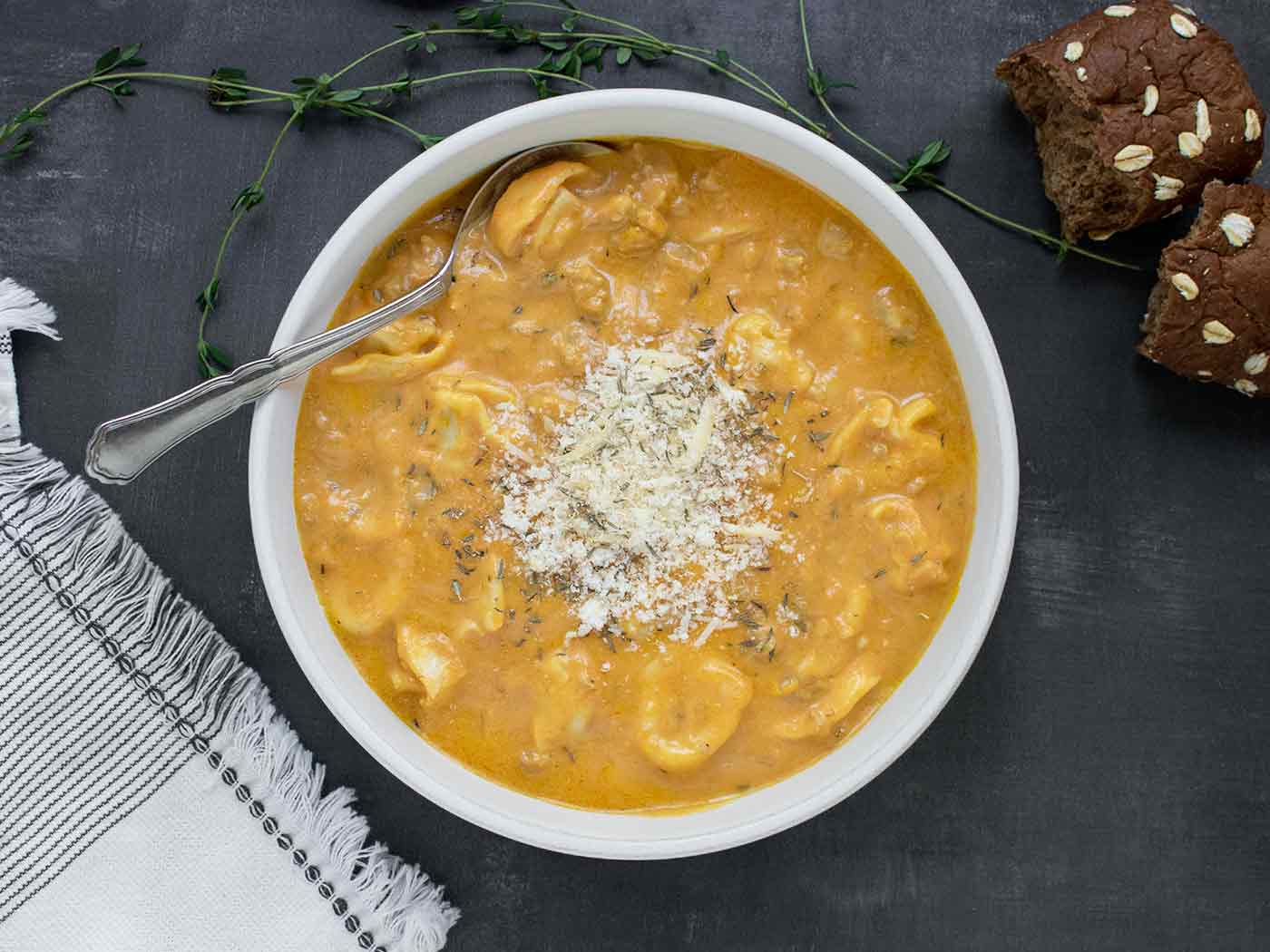 Overhead shot of a bowl of Creamy Pumpkin Sausage Tortellini Soup with a spoon and a side of dinner rolls.