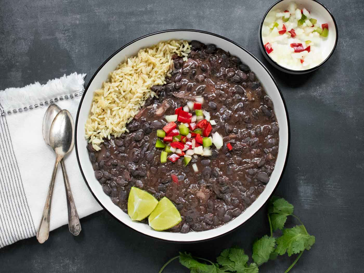 An overhead view of Cuban Black Bean Soup in a white bowl with a black rim, with rice on the side for mixing in.