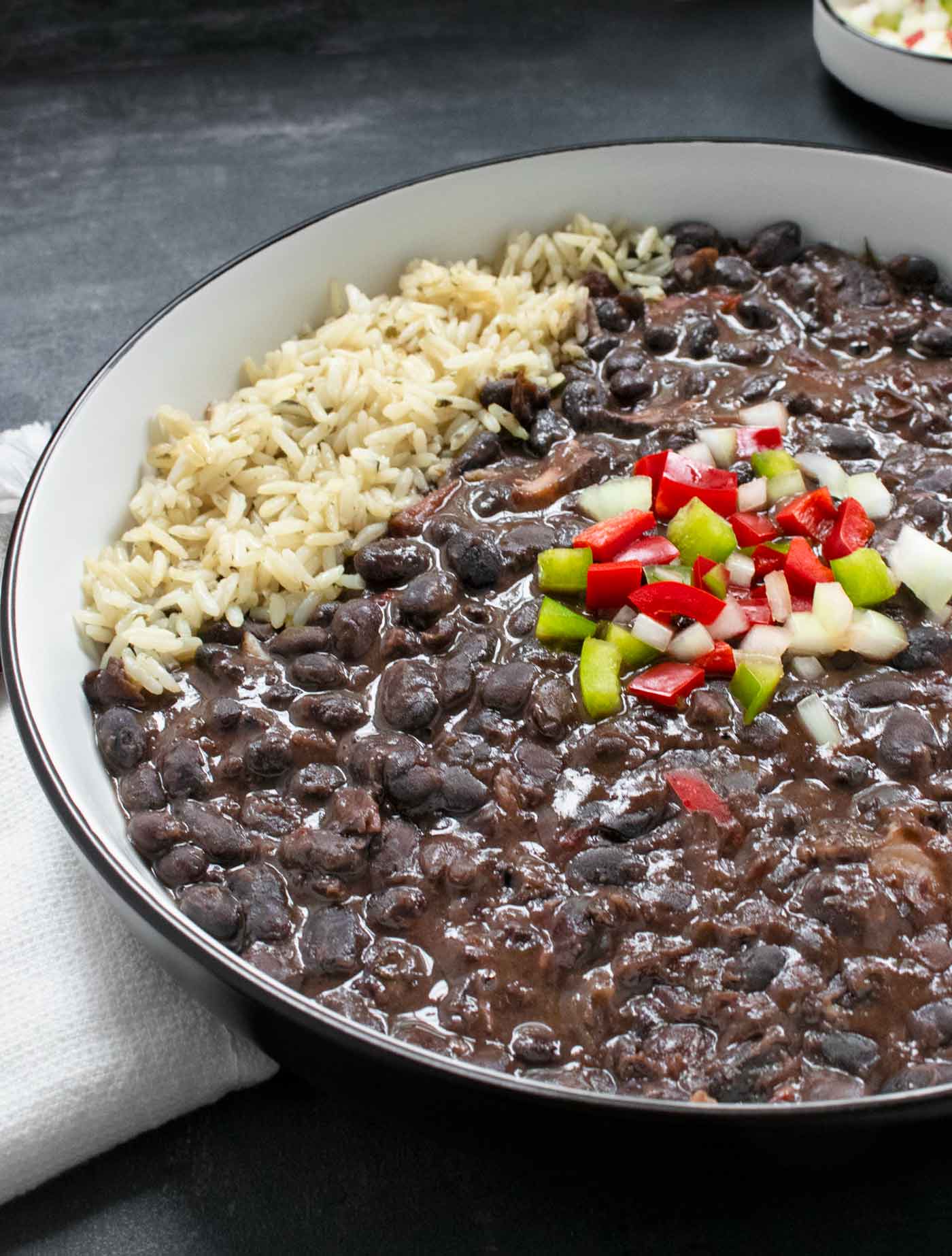 Side-angled view of Cuban Black Bean Soup in a white bowl with a black rim, with rice on the side.