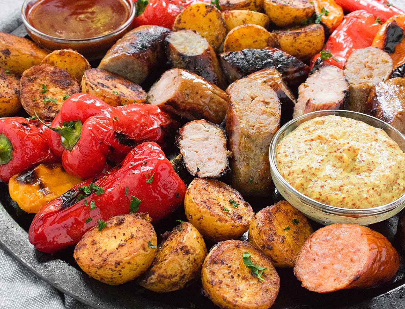 Grilled sausages, peppers, and potatoes on a platter with dipping sauces
