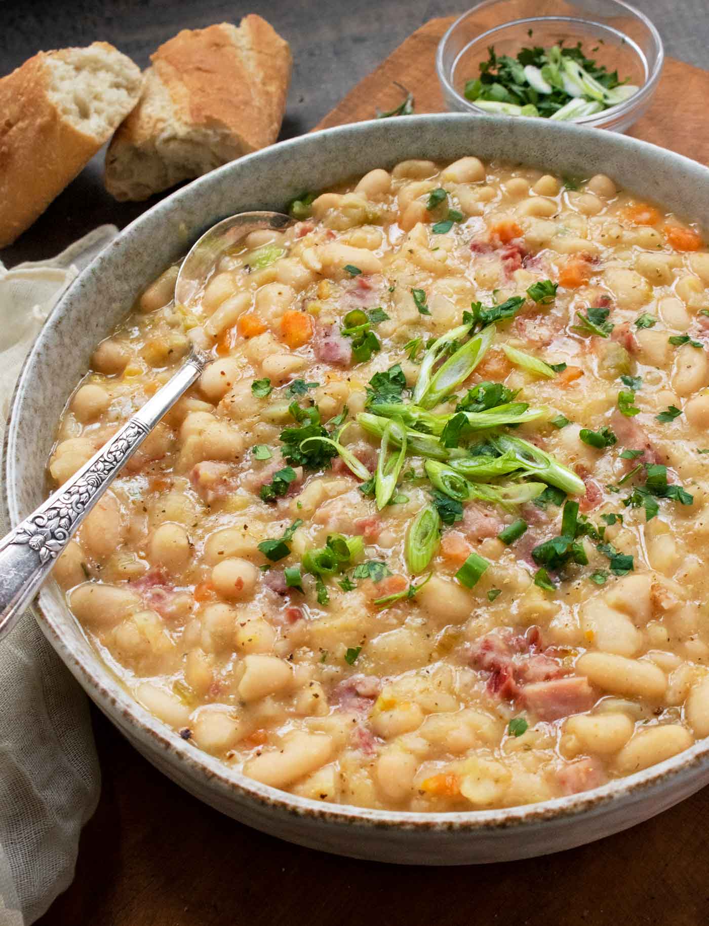 A close-up view of Ham and Bean Soup in a bowl with a spoon and slices of bread in the background.