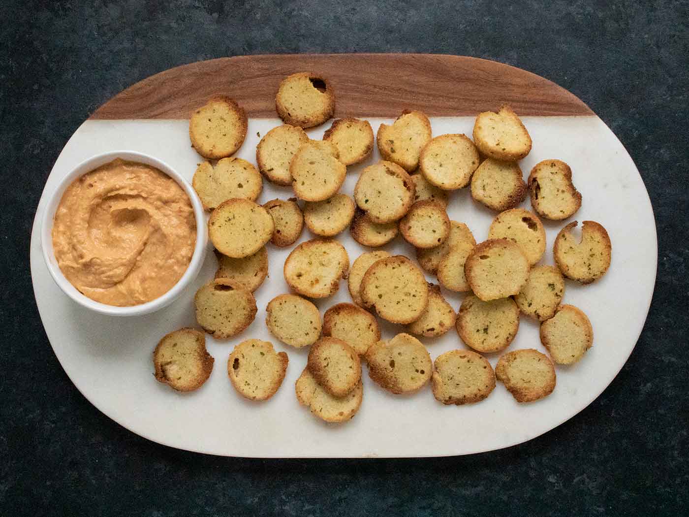 Homemade Bagel Chips on a marble serving board with a bowl of roasted red pepper hummus.