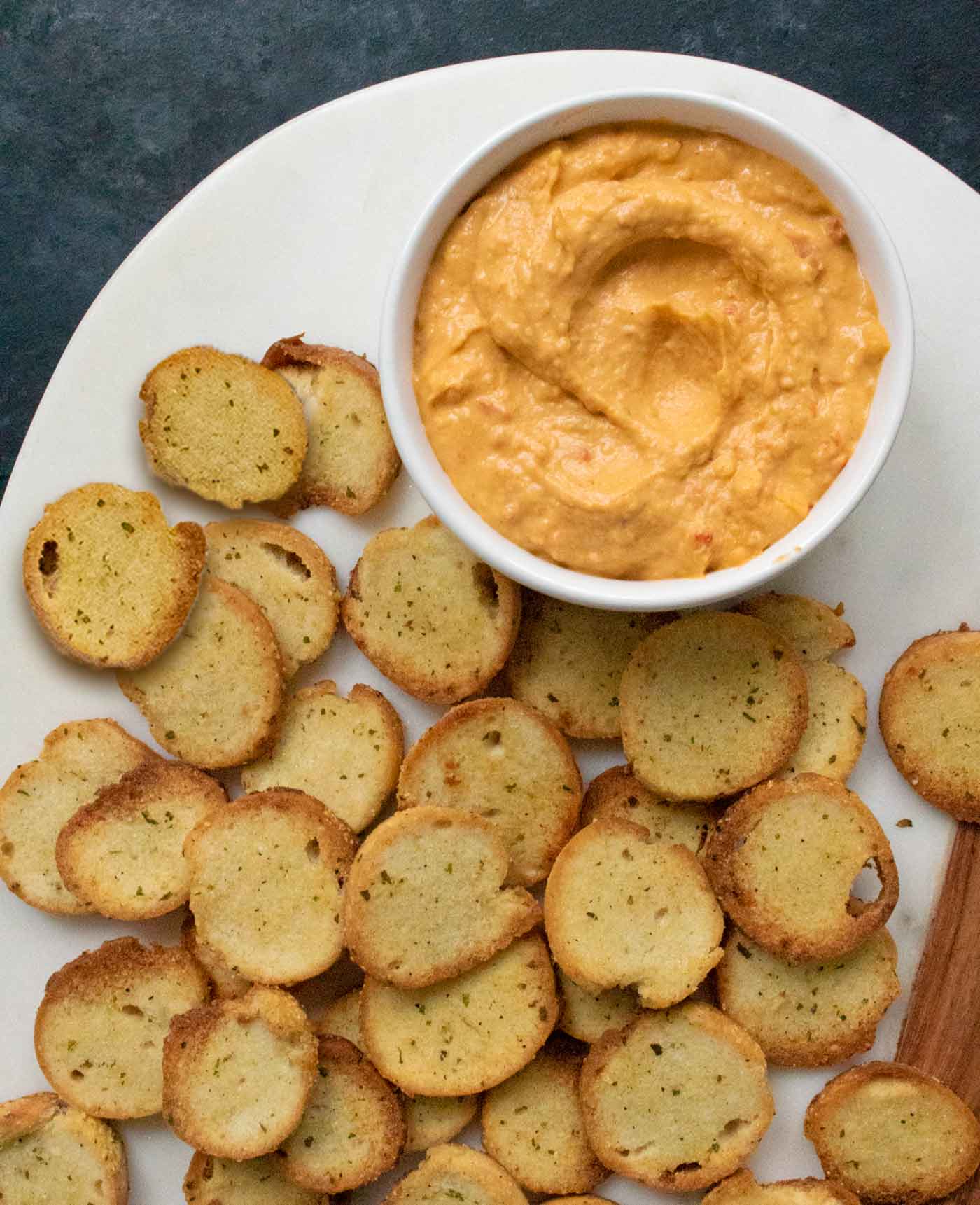 Homemade Bagel Chips and a bowl of roasted red pepper hummus on a white serving board.
