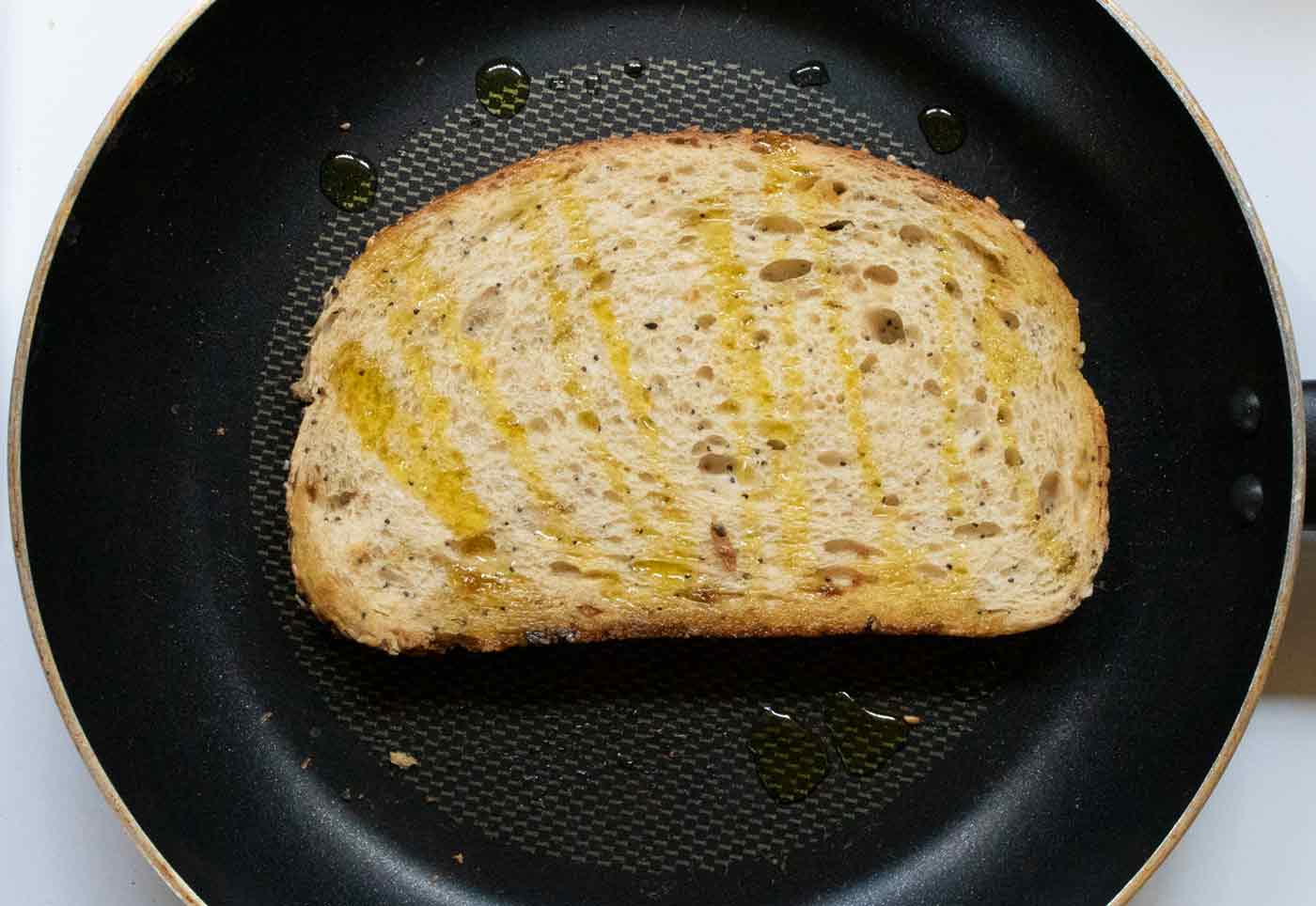 The second side of the slice of sourdough bread with olive oil drizzled over it (the underside is golden brown and the bread is ready to flip).