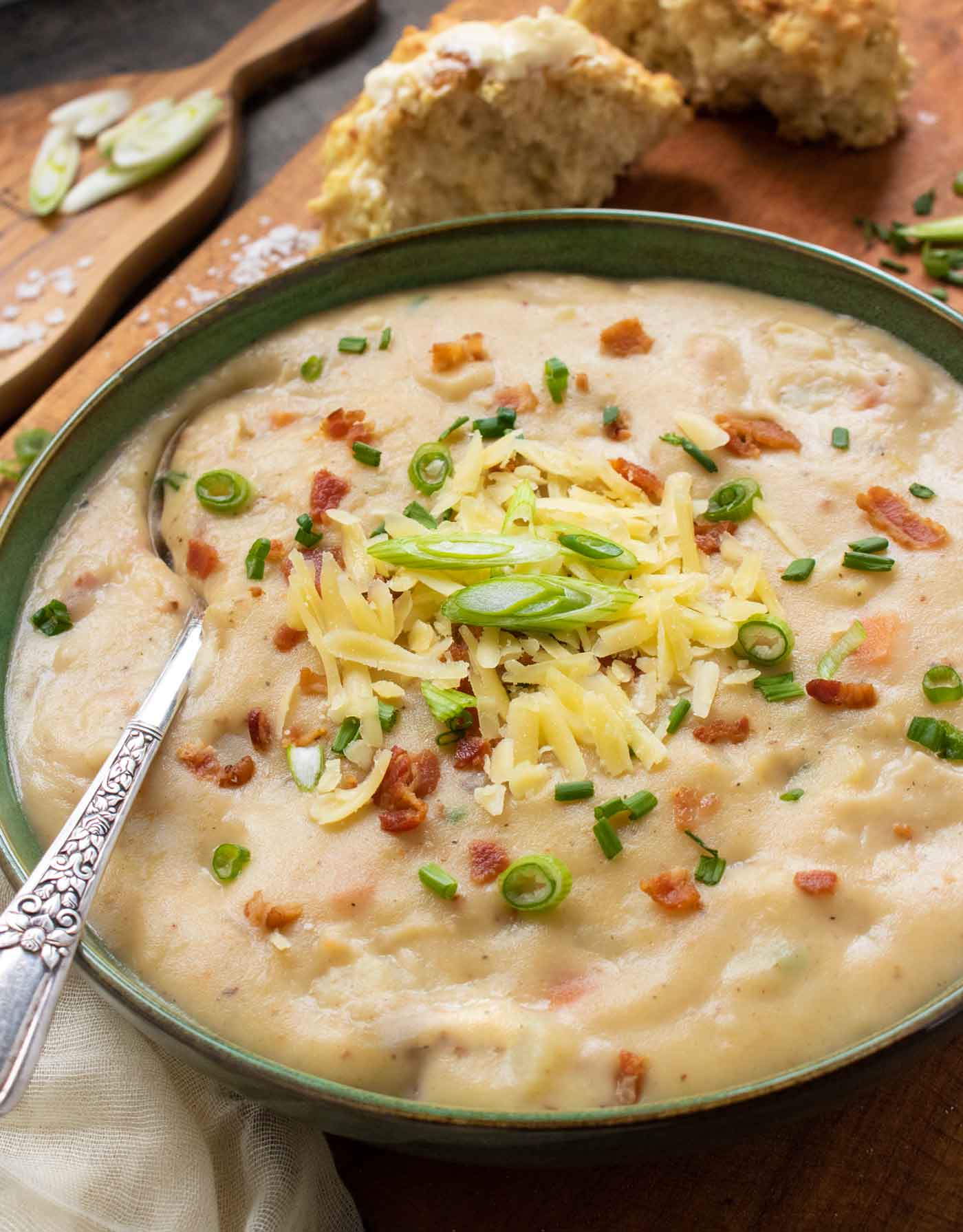 Side-angled view of Irish Potato Soup in a dark green bowl, with a spoon, and toppings.
