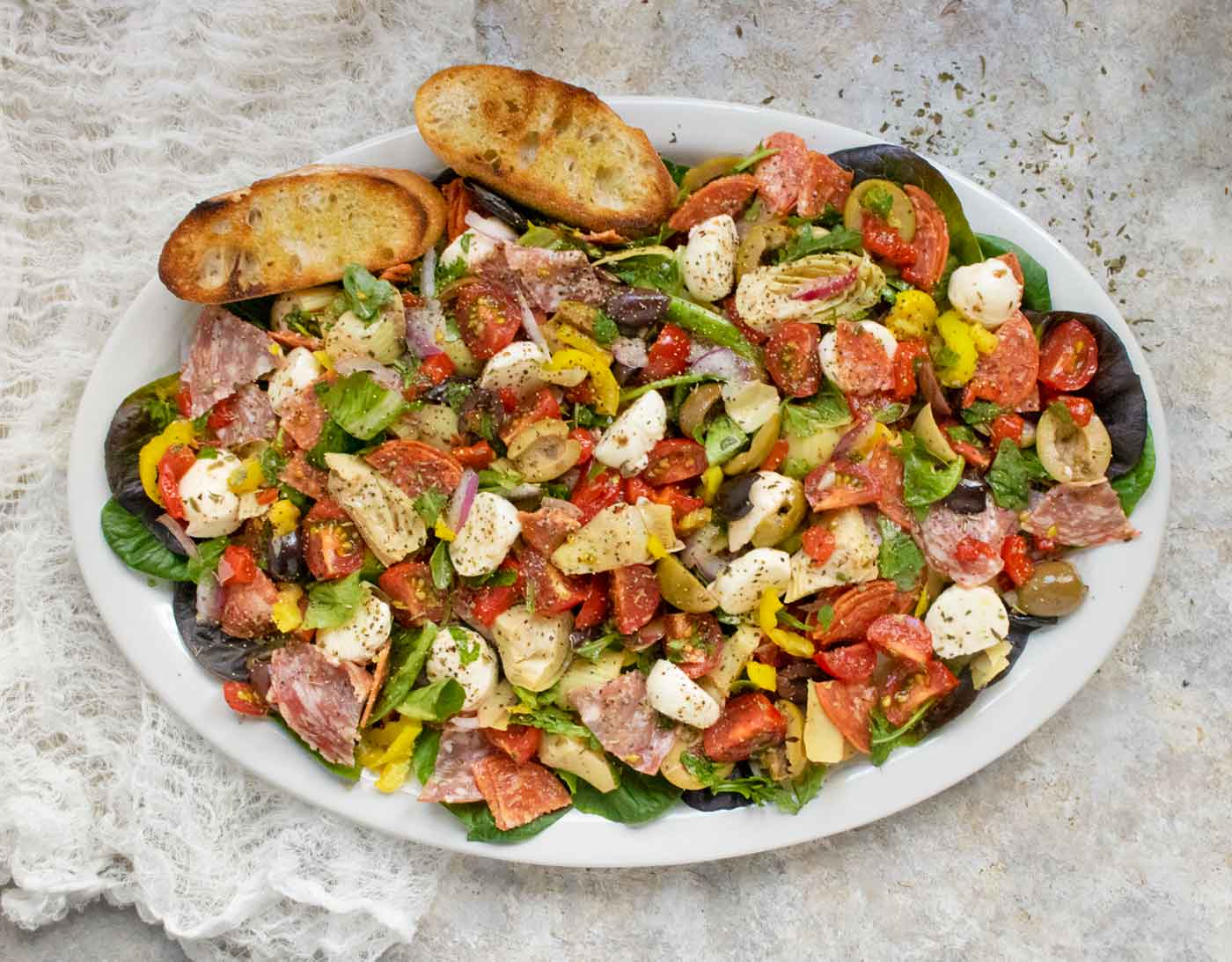 Overhead view of Italian Antipasto Salad arranged on an oval, white serving platter with slices of toasted baguette.