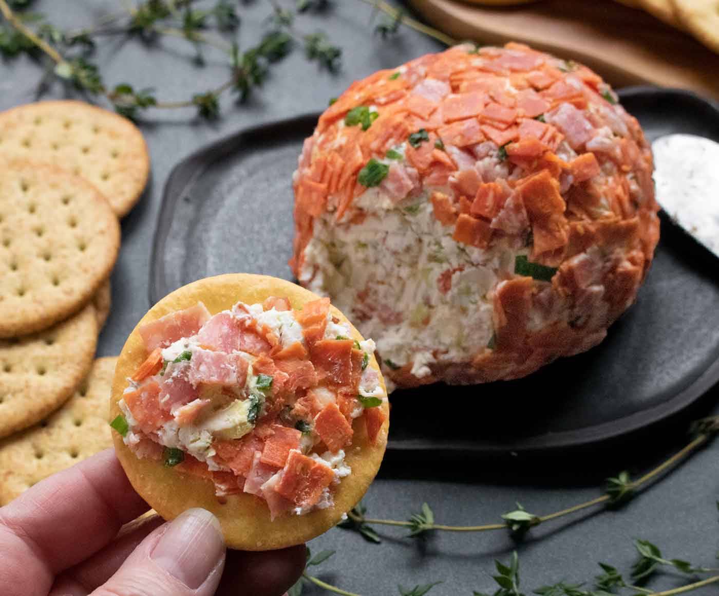 Italian Grinder Cheese Ball spread on a cracker, with the cheese ball and crackers in the background.