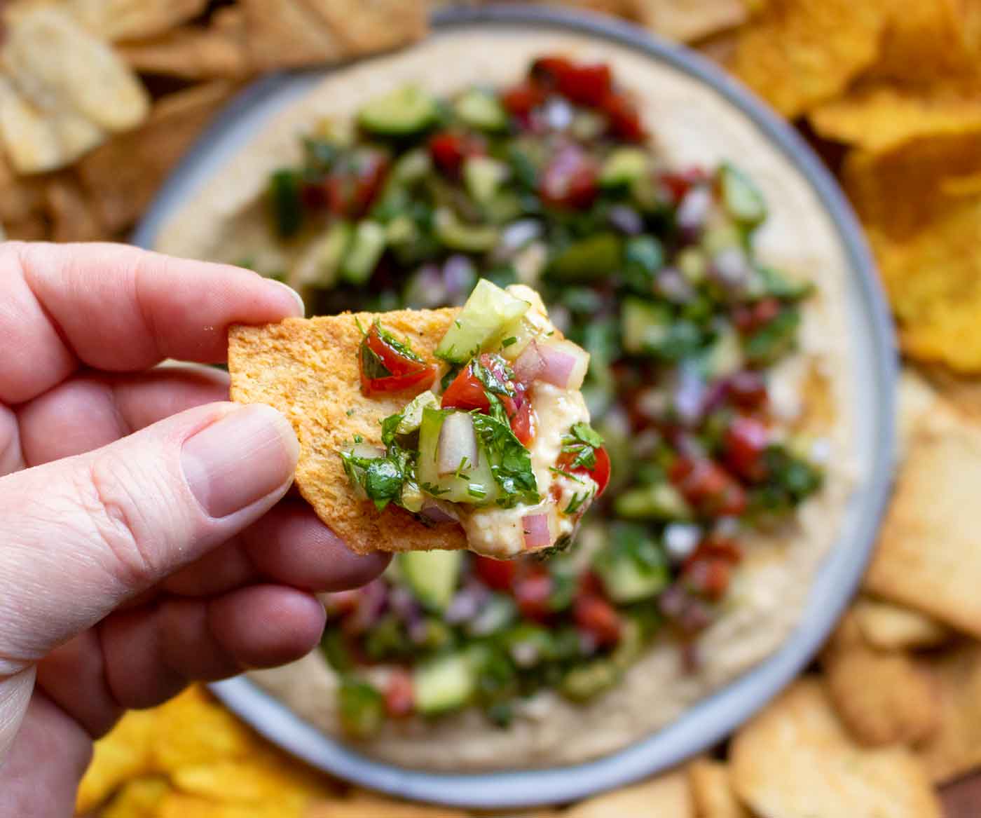 Close-up of a hand holding a chip topped with Layered Veggies & Hummus Dip.