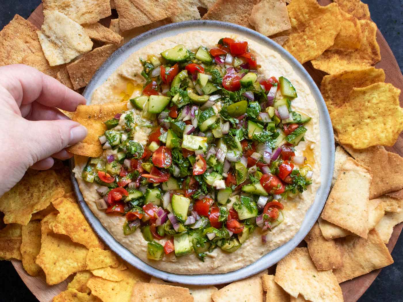 A hand scooping a chip into Layered Veggies & Hummus Dip, set on a wooden serving board with chips.