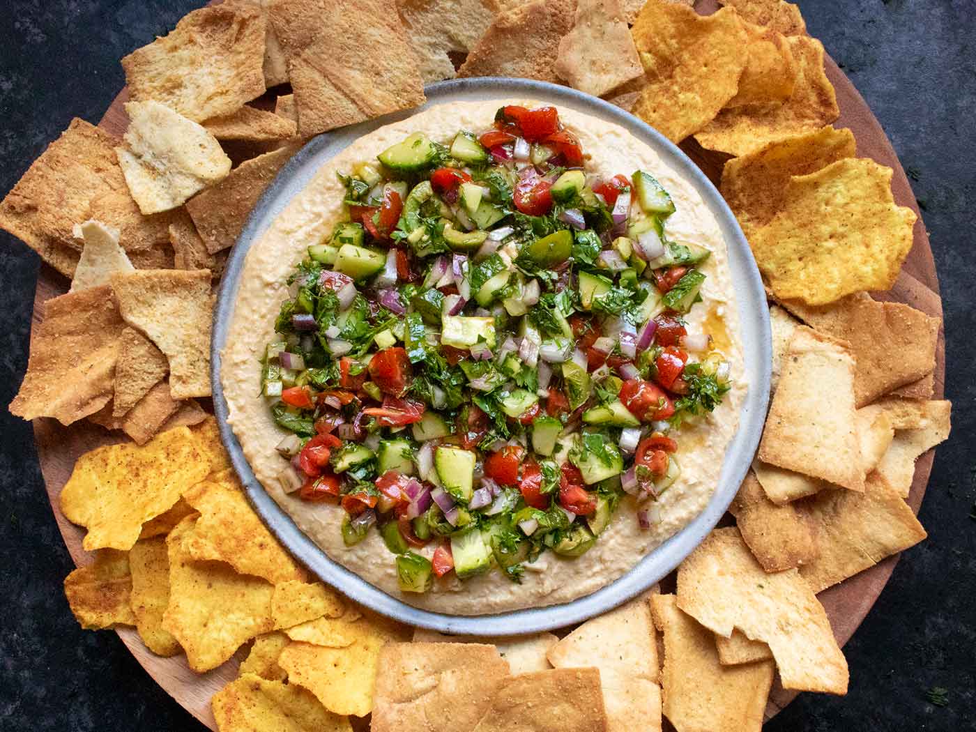 A plate of Layered Veggies & Hummus dip on a wooden serving tray with chip.