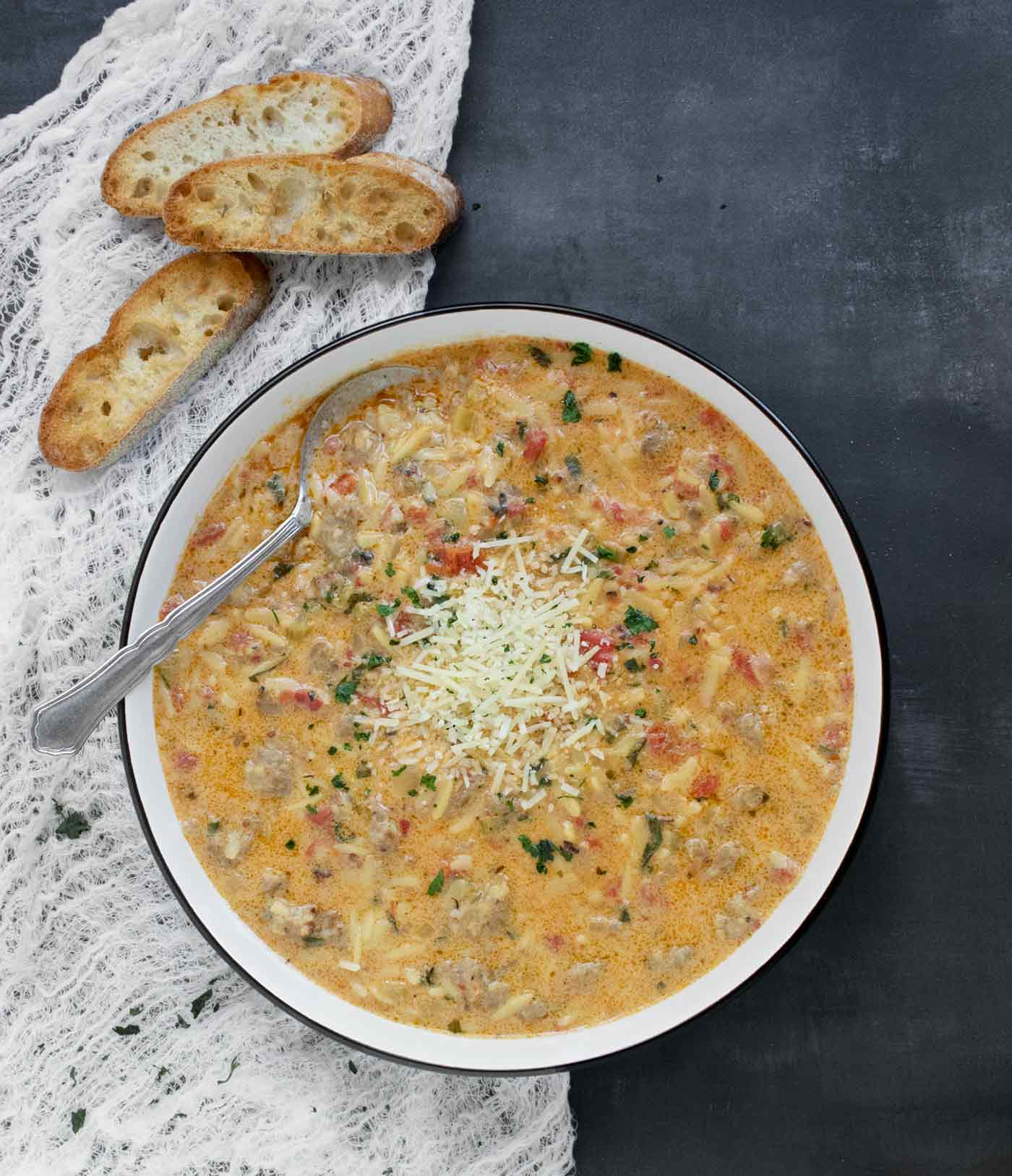 An overhead view of Marry Me Orzo Sausage Soup in a white bowl with a black rim, with a spoon and three slices from a baguette.