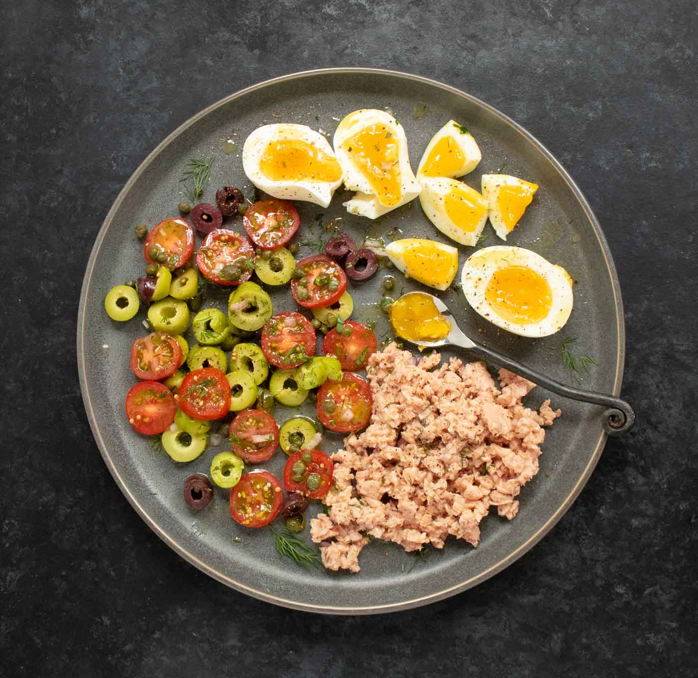 Overhead view of the main ingredients - eggs, tomatoes, olives, capers, salmon - on a dark gray plate.