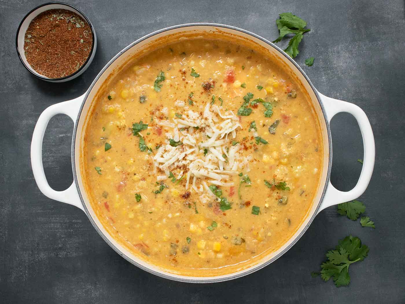 Overhead view of a white Dutch oven filled with Southwest Corn Chowder, with extra spices on the side in a small bowl.