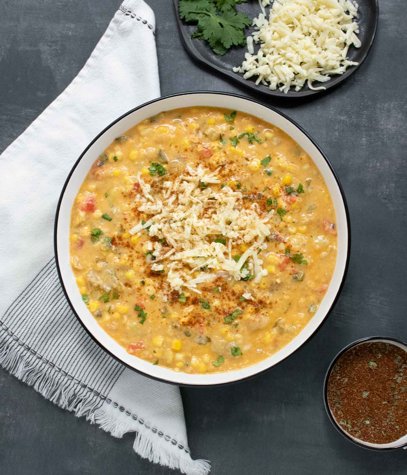 Overhead view of Southwest Corn Chowder in a white bowl with a black rim, with cheese, cilantro, and spices on the side.