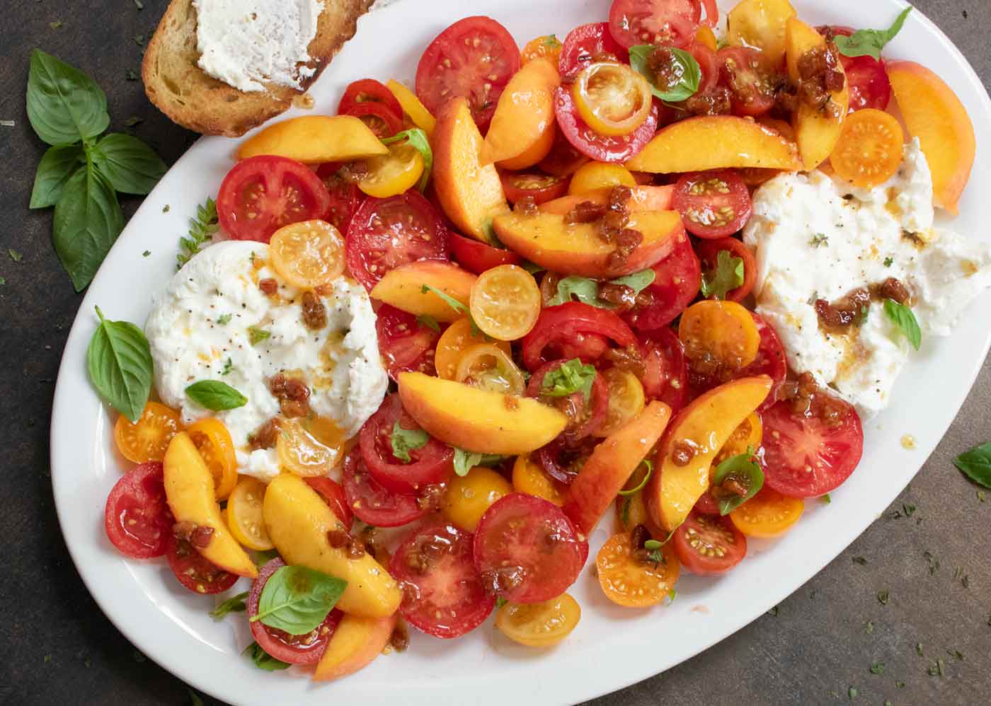 Tomato Peach Burrata Salad on a white oval platter with herbs and bread.