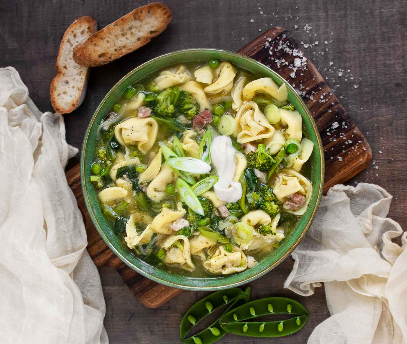 Photo from above of Tortellini Primavera Soup in a green bowl on a wooden serving board.